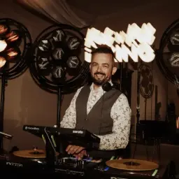 Uśmiechnięty DJ przy konsoli, gotowy do imprezy. Zastanawiasz się, ile kosztuje dj na wesele? Ten profesjonalista zapewni oprawę muzyczną.