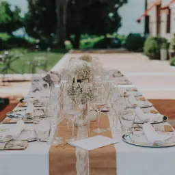 Elegancko nakryty stół na wesele z kieliszkami i serwetkami. W tle zielony ogród i budynek. Idealne miejsce na toast dobrym winem na wesele.
