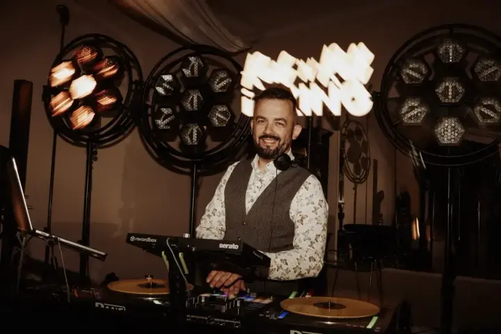 Uśmiechnięty DJ przy konsoli, gotowy do imprezy. Zastanawiasz się, ile kosztuje dj na wesele? Ten profesjonalista zapewni oprawę muzyczną.