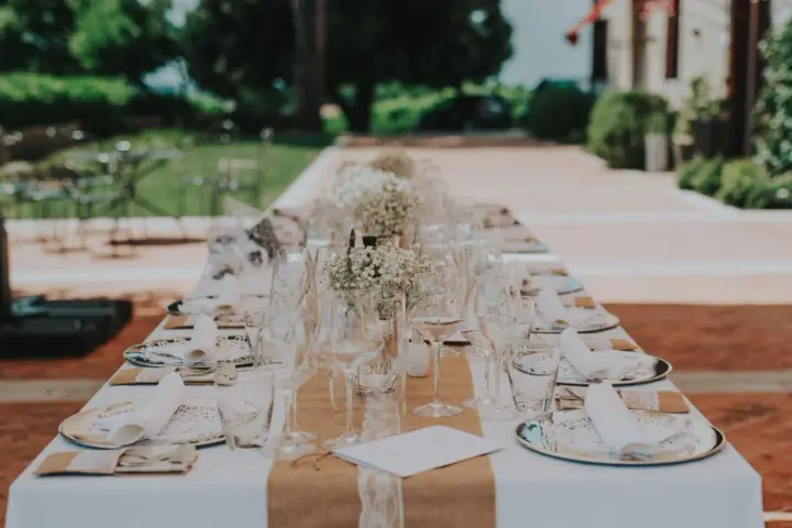 Elegancko nakryty stół na wesele z kieliszkami i serwetkami. W tle zielony ogród i budynek. Idealne miejsce na toast dobrym winem na wesele.