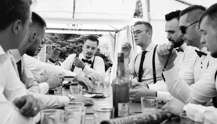 Panowie w eleganckich koszulach i szelkach świętują kawalerski pomysły przy stole z cygarami i drinkami.