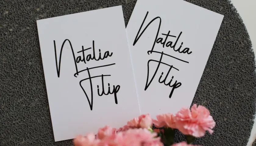 Dwie białe kartki z napisem "Natalia Filip" w ozdobnej czcionce, na tle szarej tekstury i różowych kwiatów.