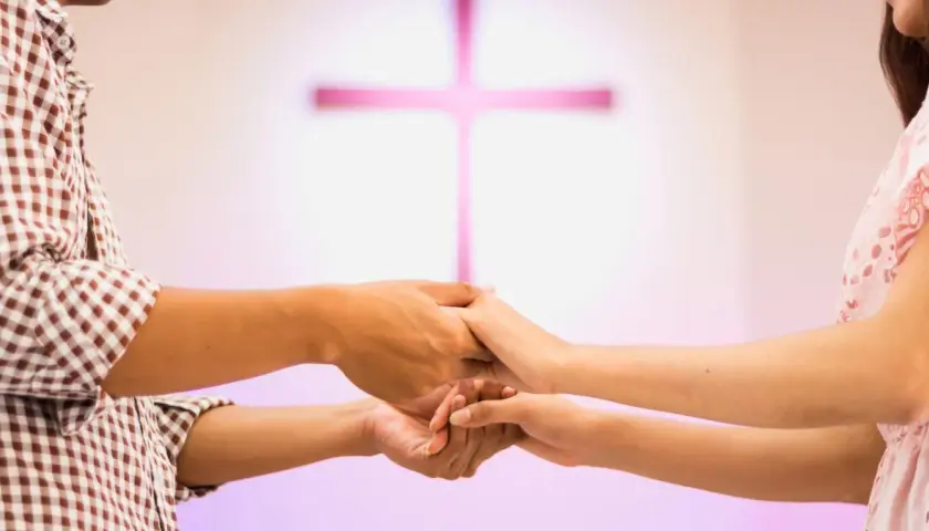 Para młoda trzyma się za ręce, przygotowując się do nauk przedmałżeńskich. W tle różowy krzyż.