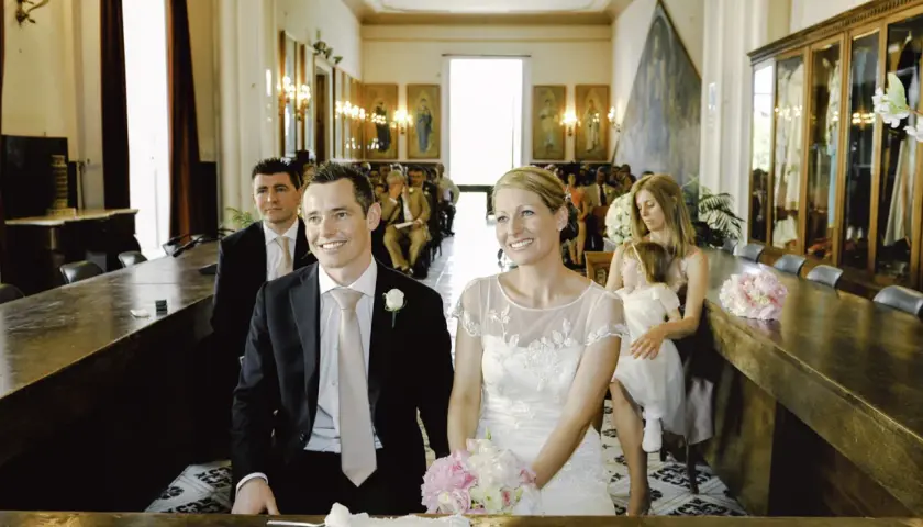 Uśmiechnięty świadek na ślubie, obok panny młodej, w eleganckim czarnym garniturze i jasnoróżowym krawacie.